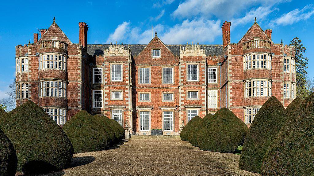 Front view of Burton Agnes Hall, an Elizabethan manor in East Yorkshire, featuring red brick walls, large mullioned windows, and two symmetrical projecting wings. The foreground shows a gravel path flanked by neatly trimmed, cone-shaped topiary bushes under a bright blue sky with scattered clouds.