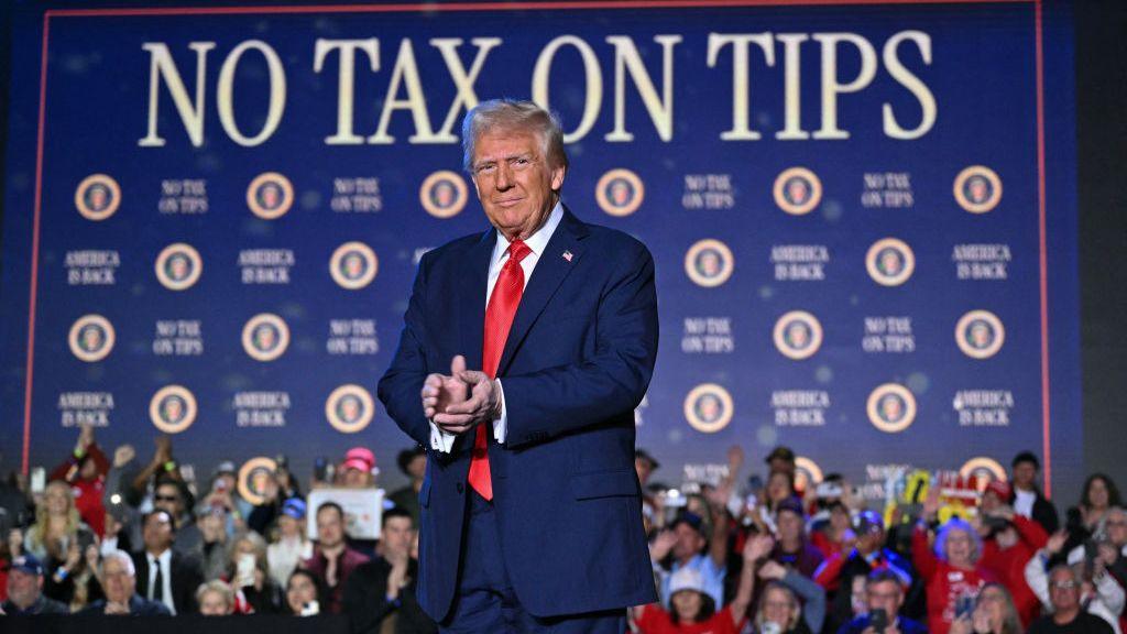 Donald Trump, wearing a blue suit, stands on stage in Las Vegas with a "No Tax on Tips" sign in the background