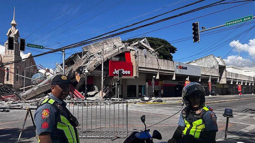 Two policemen in uniform, behind them are images damaged by an earthquake in General Santos Philippines, including a Jollibee fast food branch