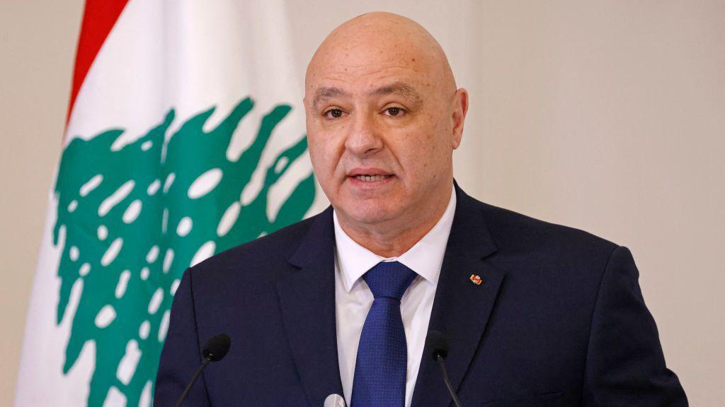 Joseph Aoun, a bald man wearing a blue suit and blue tie with white shirt stands in front of the Lebanese flag.