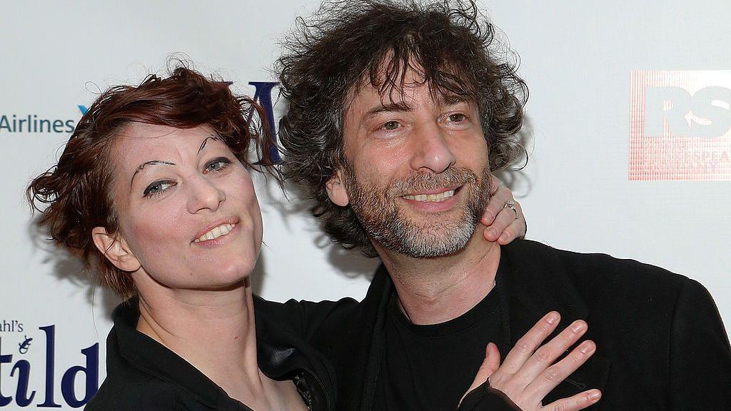 A mid shot: Palmer, a woman with tattooed eyebrows and dyed red hair, rests her hands on the chest and back of the neck of Gaiman, bearded with shaggy hair, as they pose, wearing black, at a media event