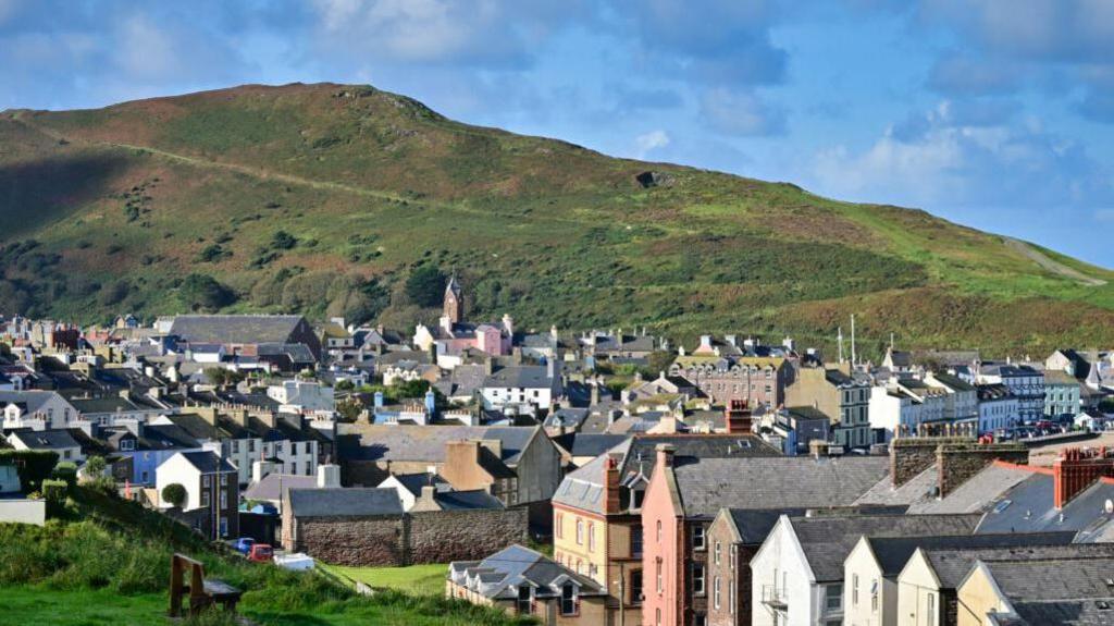 An view of the rooftops in the town of Peel with Peel Hill as a backdrop. The sky is bright blue with some clouds.