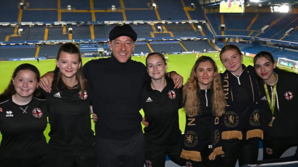 Six members of the women's team, wearing their matching kits, stand next to John Terry, who wears a black cap and jumper. They all smile at the camera with their arms around each other, at Stamford Bridge football ground. Blue seats and the football pitch are seen behind them.