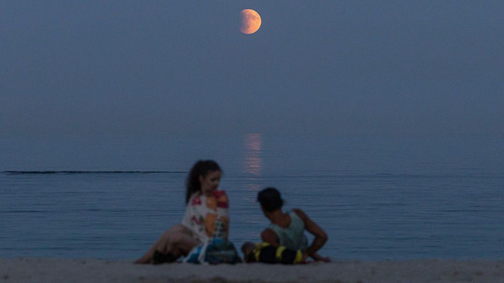 Two people laying on the beach in front of the Blood Moon which is reflected in the sea