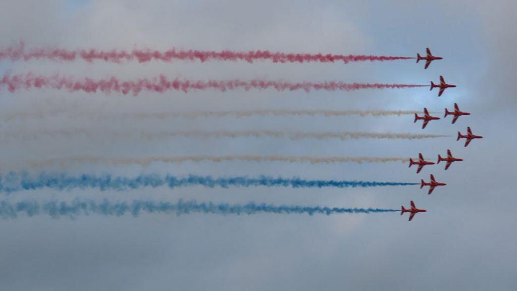 The Red Arrows flying in Guernsey. There are nine aircraft flying in the sky with red orange and blue smoke. 