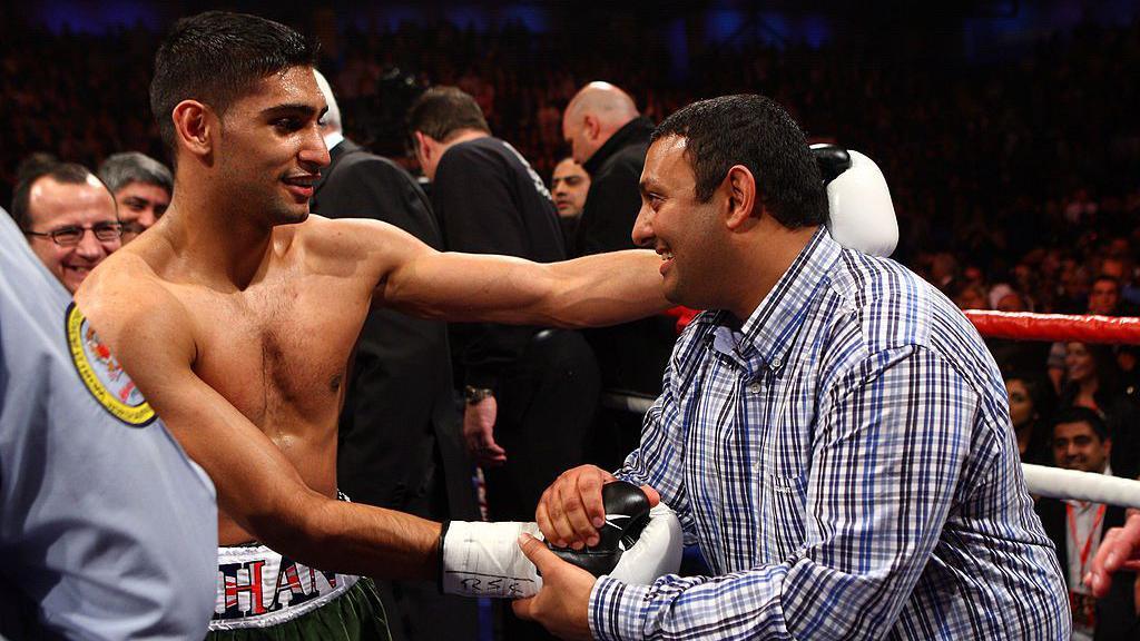 Two men shake hands in a boxing ring. One, Amir Khan, is wearing gloves and shorts at the end of his fight. The other, Naseem Hamed, is wearing a blue chequered shirt. Both men are smiling.
