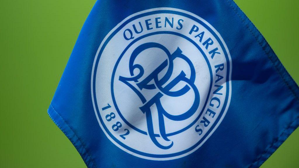 A file image showing a small blue corner flag bearing the logo of QPR in white. Green grass can be seen in the out-of-focus background