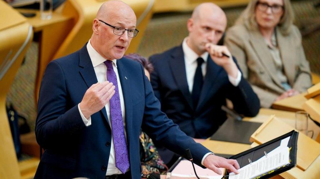 First Minister John Swinney stands in Holyrood's chamber turning the page of a black folder with ministers sitting in the background