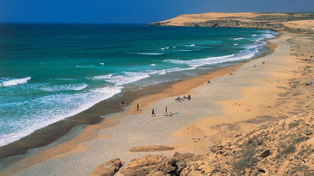 A sandy beach which meets a blue and green sea. The outline of people are visible on the beach