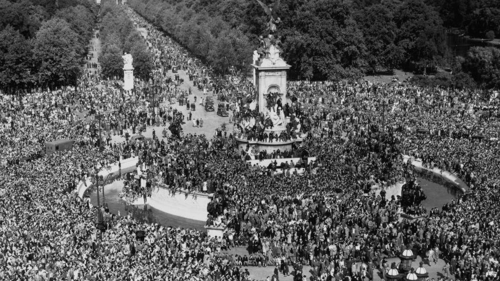 A high-angle black-and-white view of crowds of people celebrating VJ Day, some climbing on the Victoria Memorial, on The Mall outside Buckingham Palace in Westminster, London.