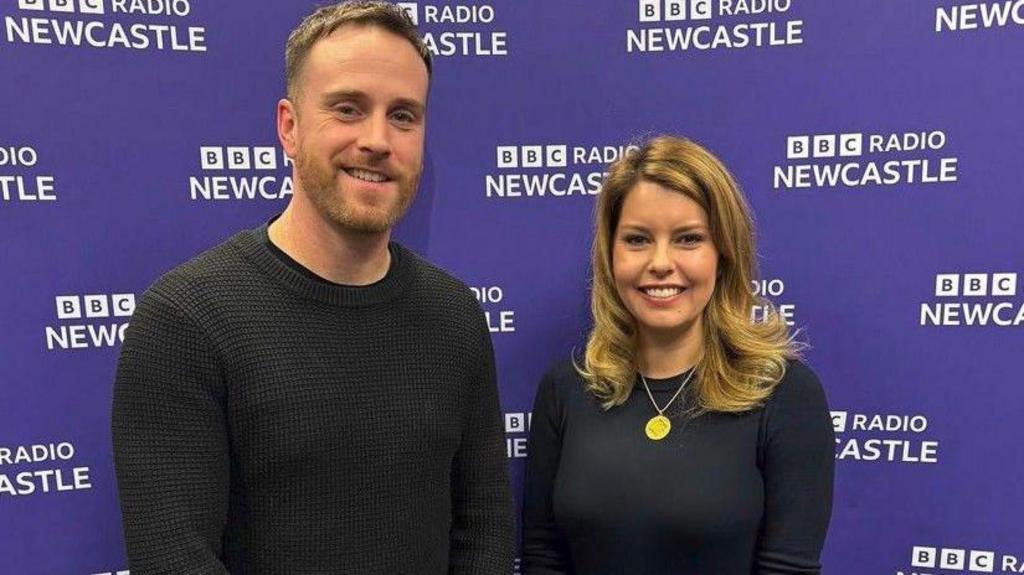 Breakfast Presenter Matt Bailey and North East Mayor Kim McGuiness are standing in front of a purple background. The Radio Newcastle logo is on the wall in white several times. They are both smiling. 