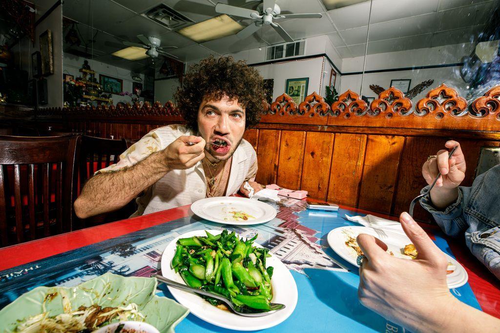 Benny Blanco sitting in a restaurant putting a spoonful of food into his mouth. A vegetable dish is on the table in front of him.