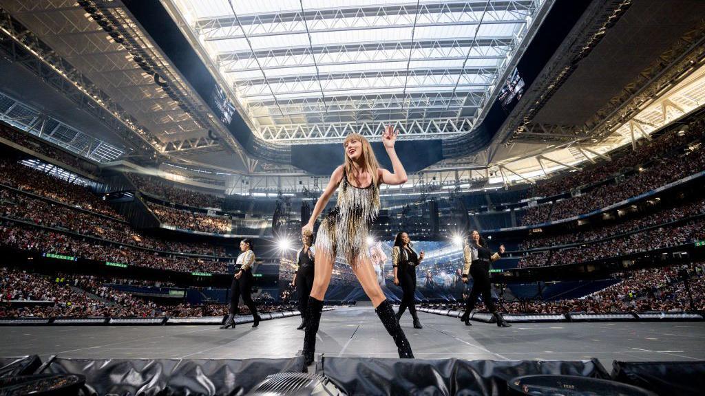Taylor Swift stands dancing at front of stage with some backing dancers behind her surrounded by thouands in the crowds at the stadium