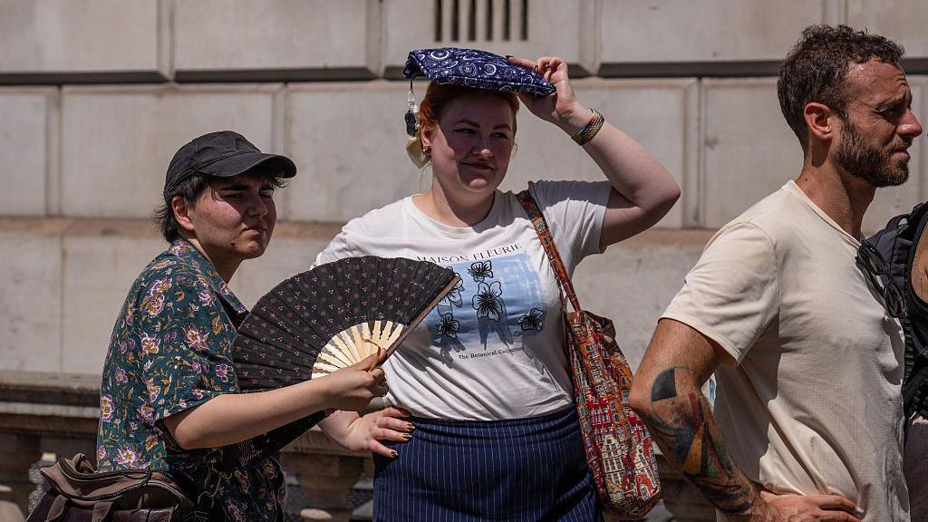 A woman fans herself as another shelters from the sun as they wait to take a picture in front of a phone box in Parliament Square. In front of them is a man wearing a beige top.