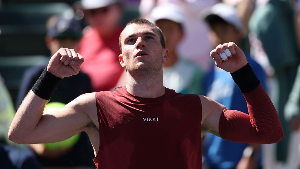 Jack Draper raises his arms to celebrate victory. He has short hair and is wearing a marron vest and a maroon sleeve on his left arm. Members of the crowd can be seen in the background.