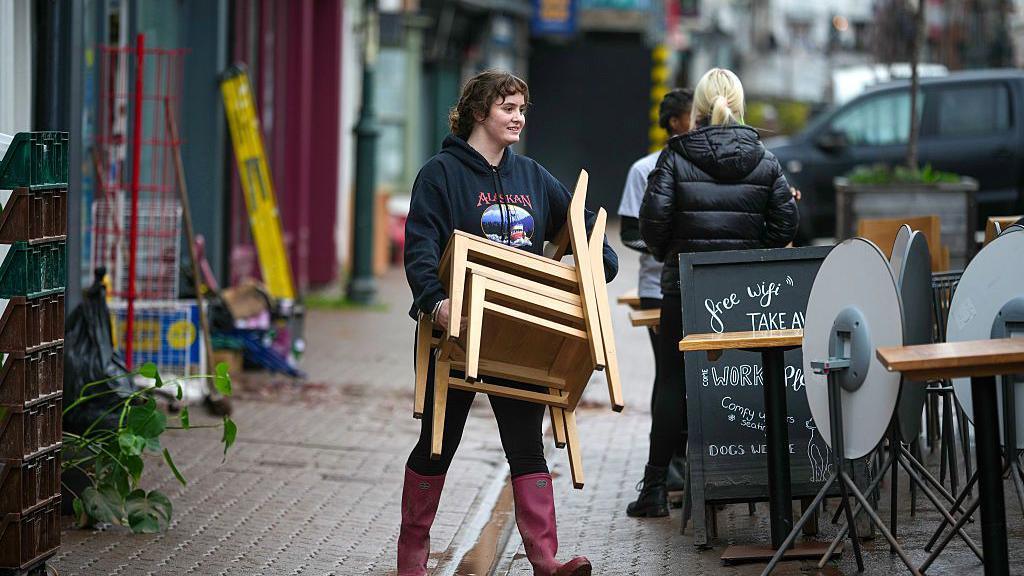 Merch yn cario cadeiriau ar y stryd yn Nhrefynwy, mae hi'n gwisgo esgidiau glaw ac yn helpu i symud y dodrefn fel rhan o'r broses glanhau.