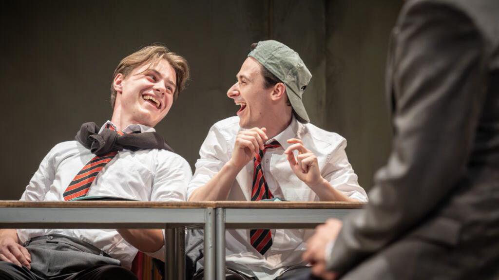 Two actors sit behind a desk. Both are laughing while looking at one another. They are wearing school uniforms. 