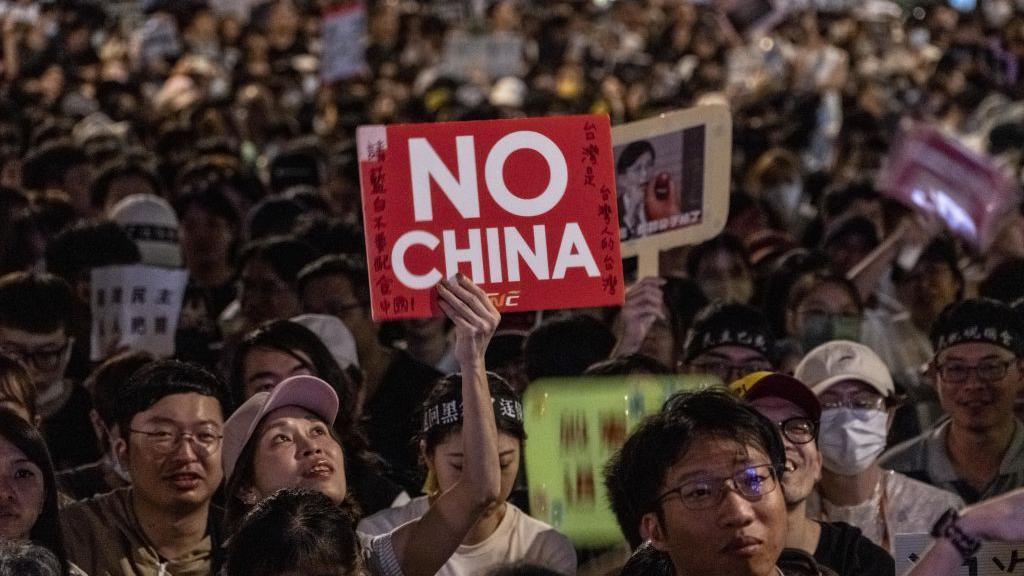 At a night-time rally, a woman in a pink cap holds up a red and white sign that says in English "No China". Surrounding her are other protesters holding up other signs as well.