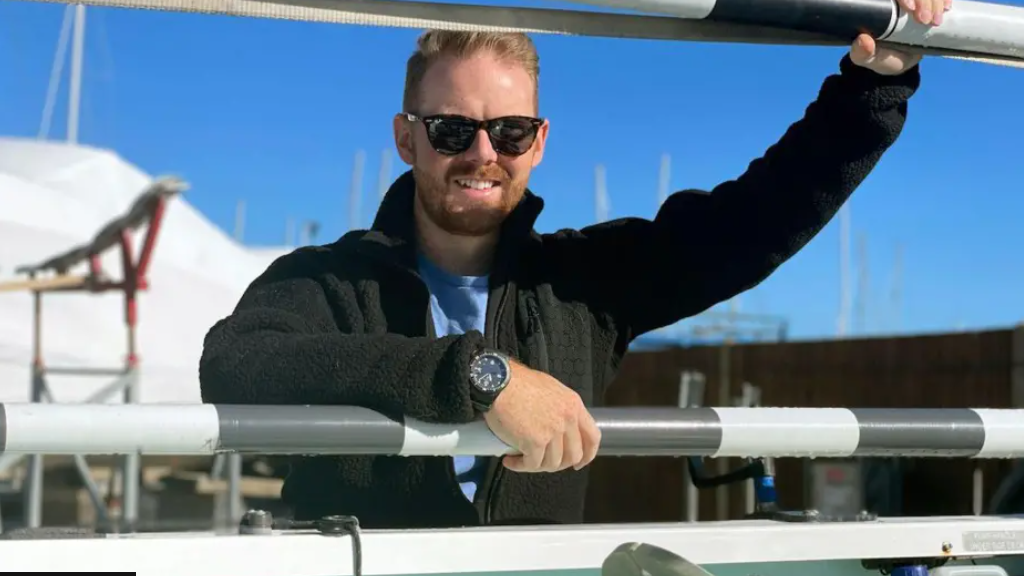 Kyle Meredith stands facing the camera. He is wearing sunglasses and a black fleece and a blue shirt. He is leaning on a black and white fence