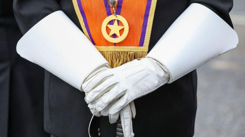 A person in white gloves holding the top of a sword. The person is wearing an orange sash and dark suit