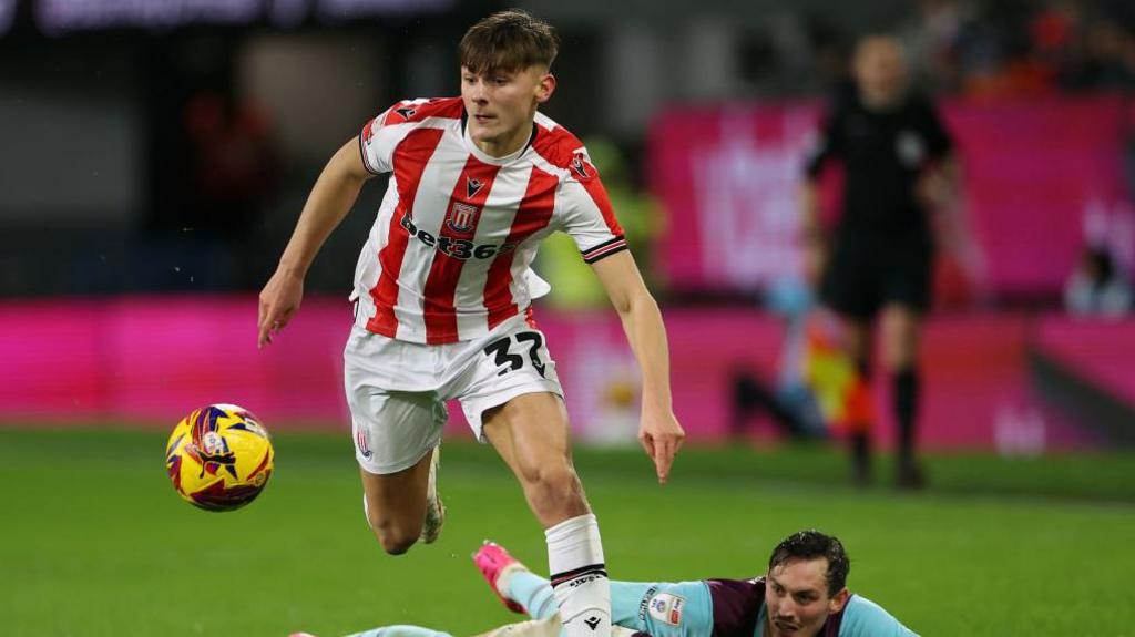Emre Tezgel shown winning a tackle and pursuing the ball in his red and white striped Stoke kit, with the opposition player left on the ground