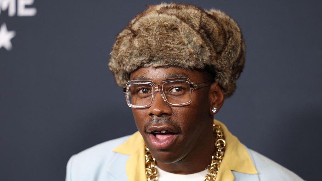 Tyler, the Creator poses wearing a furry hat, blue blazer, yellow shirt and gold chain.