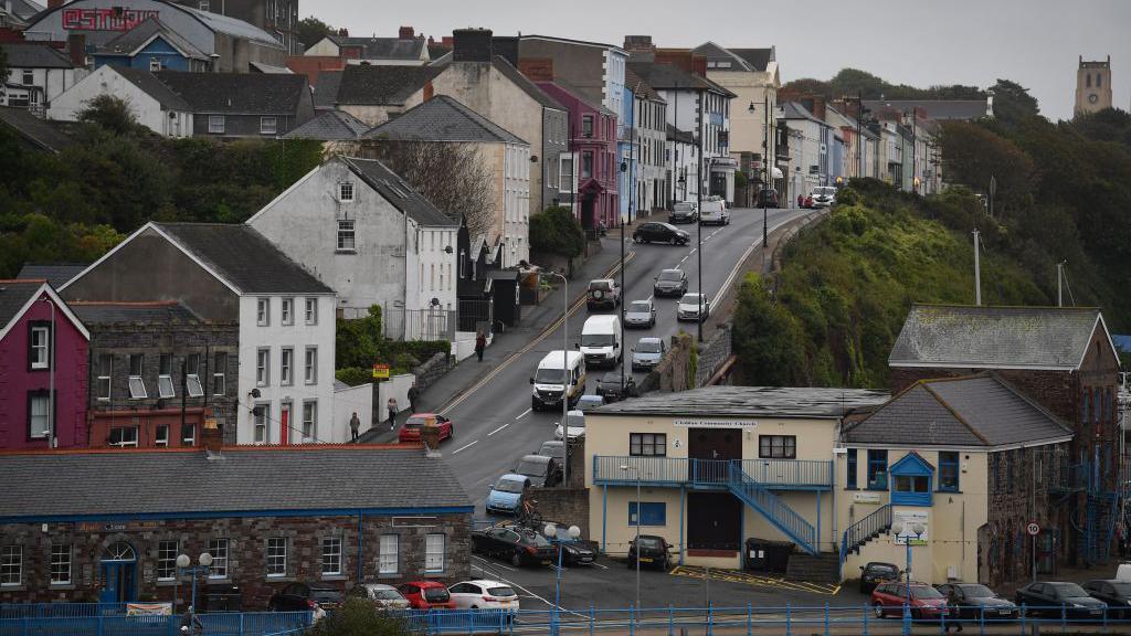 a main road in Mildford Haven along the sea side. There is a low stone building along the front beside a larger stone building with yellow rendering on one side. 