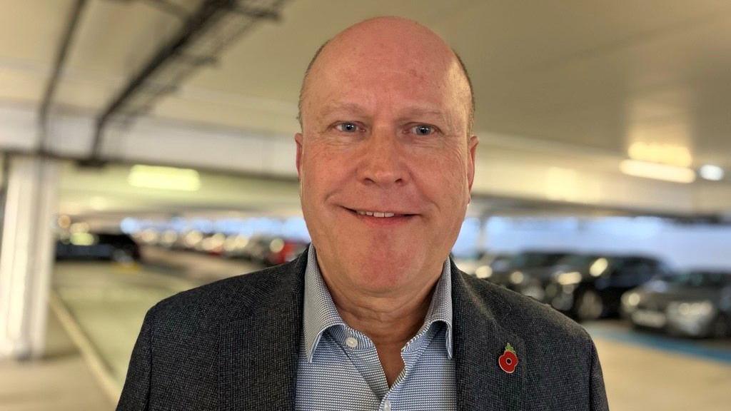 Steve is wearing a dark blue blazer and a light blue collar shirt with a red poppy.