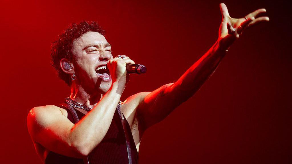 Olly Alexander stands on-stage at Manchester Pride, singing emphatically with a microphone held to his mouth in one hand and reaching out towards the audience with the other. He has a black vest on and has a large earring in his ear. The stage lighting behind him is dark red.