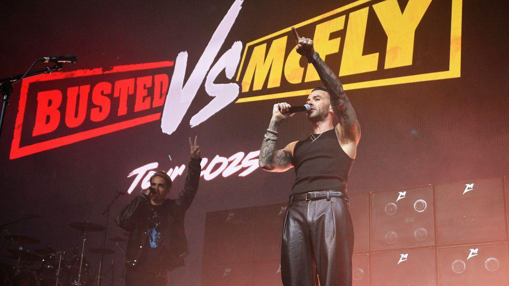 Charlie Simpson and Matt Willis from Busted stand on a stage, both with their right arms in the air. They stand in front of a screen that says "Busted VS McFly Tour 2025".