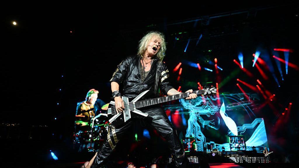 Joe Elliott is playing guitar on stage while singing. He is dressed in all black leather and has medium length white hair. The stage is dark with some bright blue and red lights shining across it.