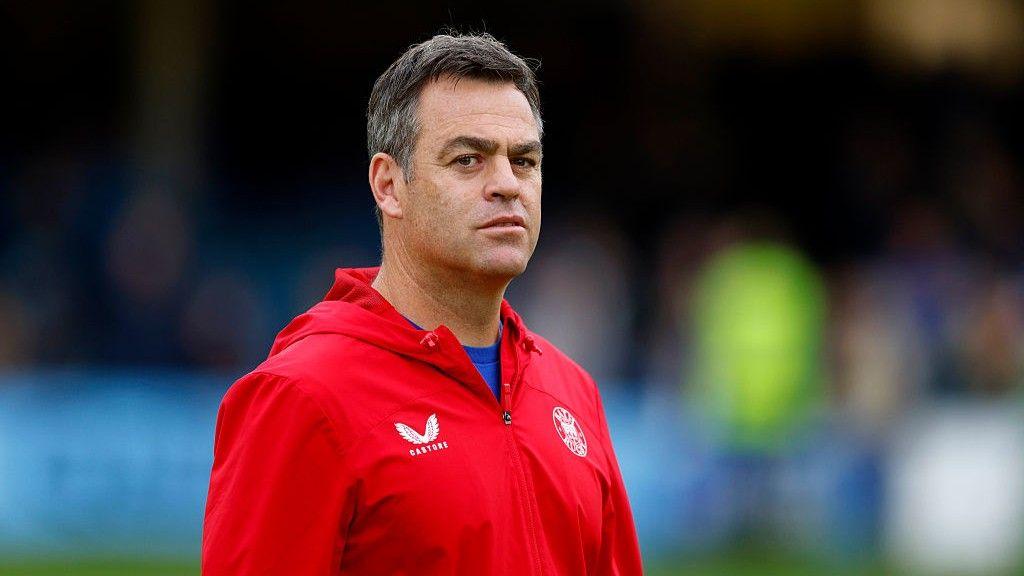 Bath director of rugby Johann van Graan looks on before a Prem Rugby fixture. Van Graan is wearing a red top, which features the logo of the Bath rugby team.