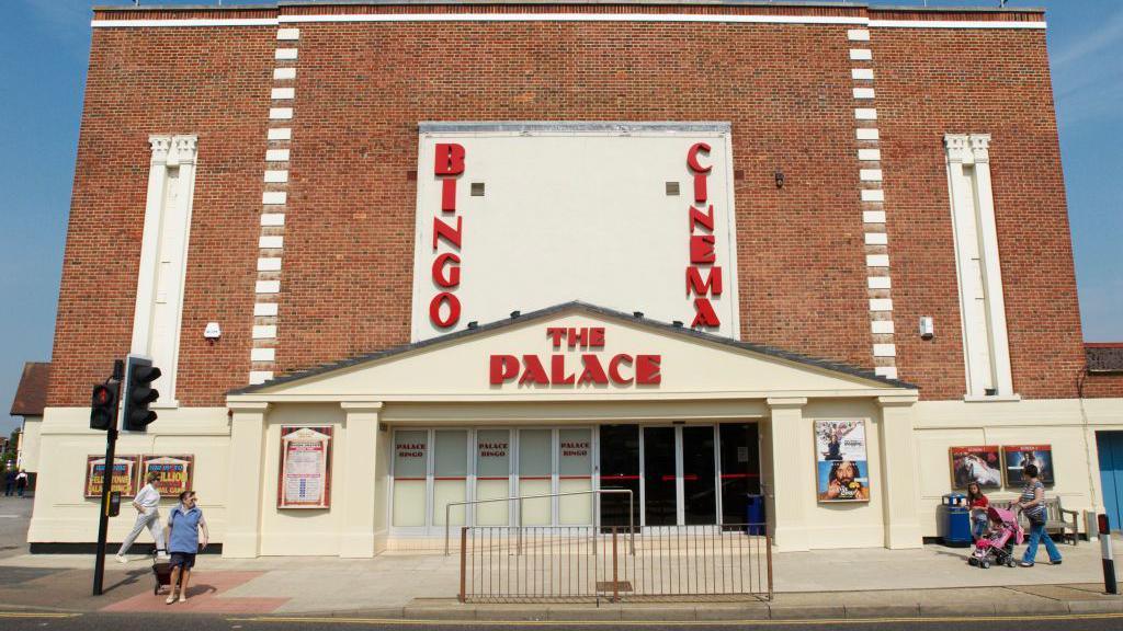 A view of the entrance into the Palace Cinema in Felixstowe. Residents walk on the pavement outside of it. The building is large and made of red bricks.