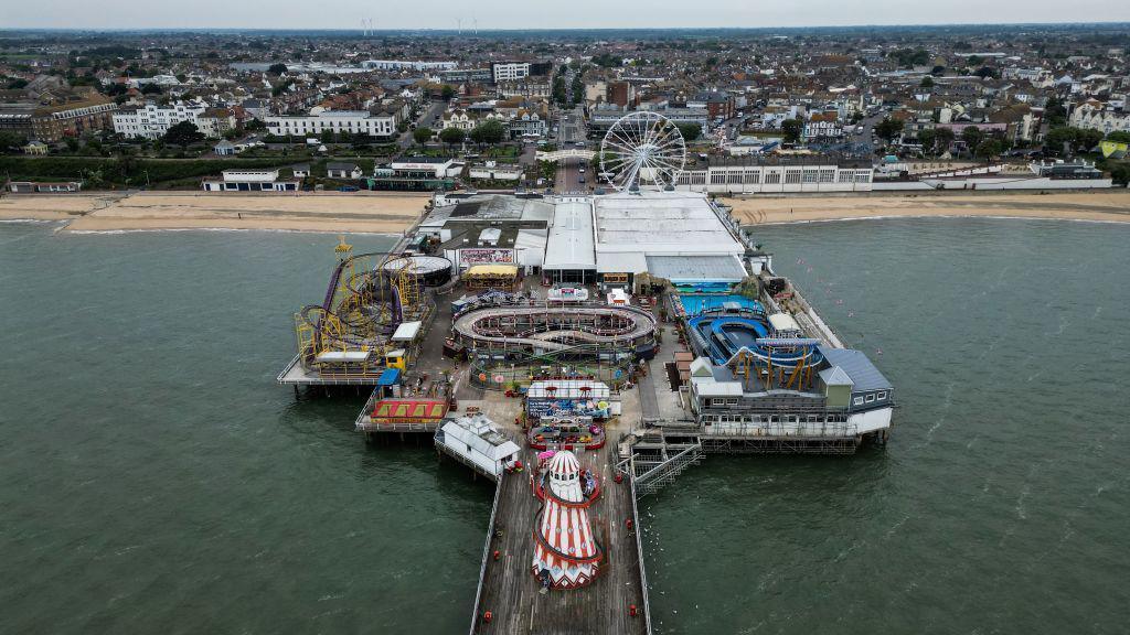 The pier at Clacton from above. The helter skelter and various other attractions are visible, including the ferris wheel. In the foreground is the beach and buildings near the shore.