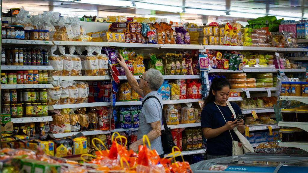 An elderly Chinese man browses through shelves of packaged bread at a supermarket in Singapore, while a woman next to him checks her phone. On the shelves you can see different types of breads, packaged cakes, and jars of food.
