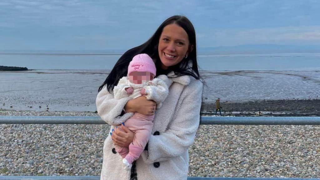 Kiena Dawes, who has long, straight black hair and wears a furry white coat, smiles at the camera while holding her baby daughter, who wears a pink hat with the words 'Mummy's Girl' stitched onto it. She is standing against railings in front of a pebbly beach.