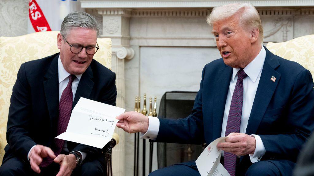 Donald Trump, seated on the right, speaks while holding up a signed document during a formal meeting. Seated next to him on the left is Sir Keir Starmer, wearing a dark suit and red tie. They are in a room with cream-colored armchairs and a white fireplace in the background.