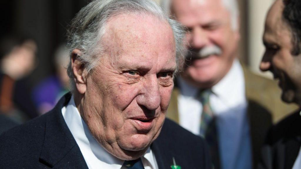 Frederick Forsyth is seen in older age in a head and shoulders portrait. He is in a crowd and looking at someone off camera.
