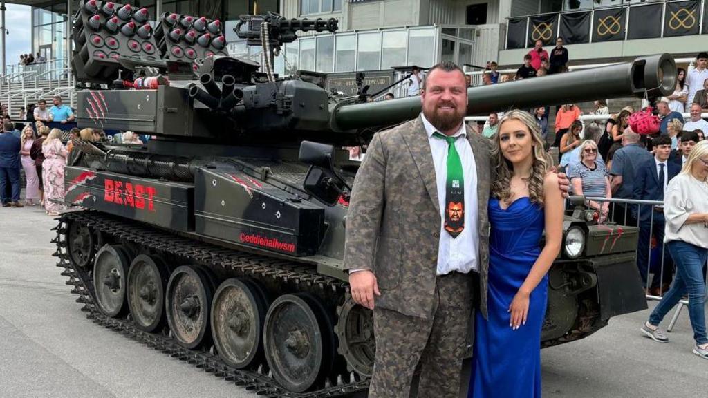 Eddie Hall is dressed in a camouflage print suit smiles as he stands in front of the tank with Molly Goodwin, who is wearing bright blue prom dress