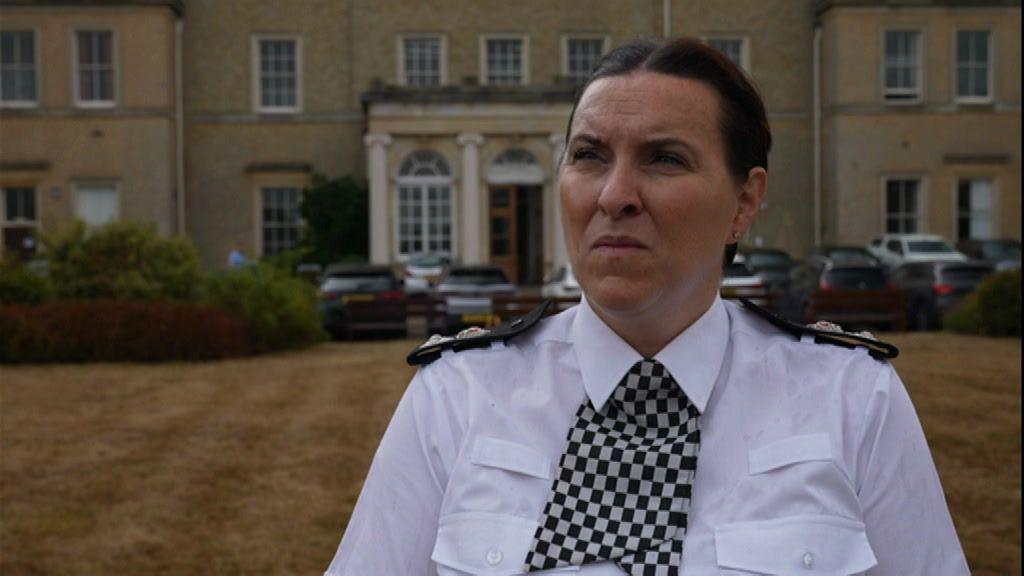 a woman with brown hair tied back. She is wearing a white shirt and a black and white checkered tie. She has police badges on both of her shoulders