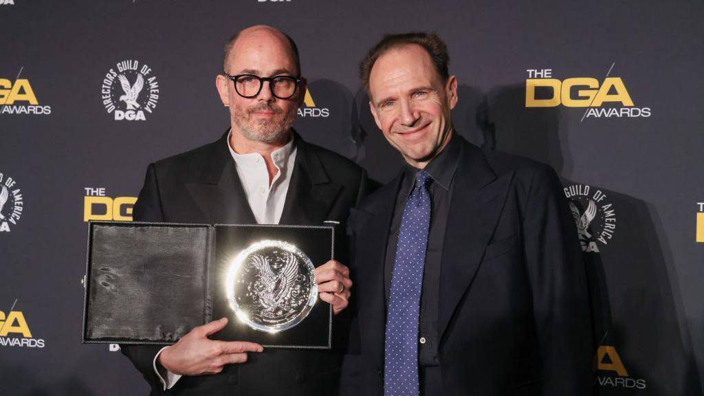 Ralph Fiennes and Edward Berger pose for a camera on a red carpet. On the right, Fiennes is wearing a dark suit and blue tie. On the left Berger is wearing a dark suit and white open neck shirt. 