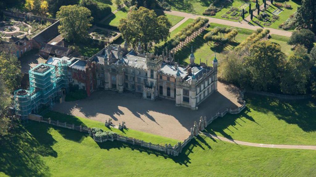 Aerial view of a huge mansion with towers surrounded by landscaped gardens and trees.