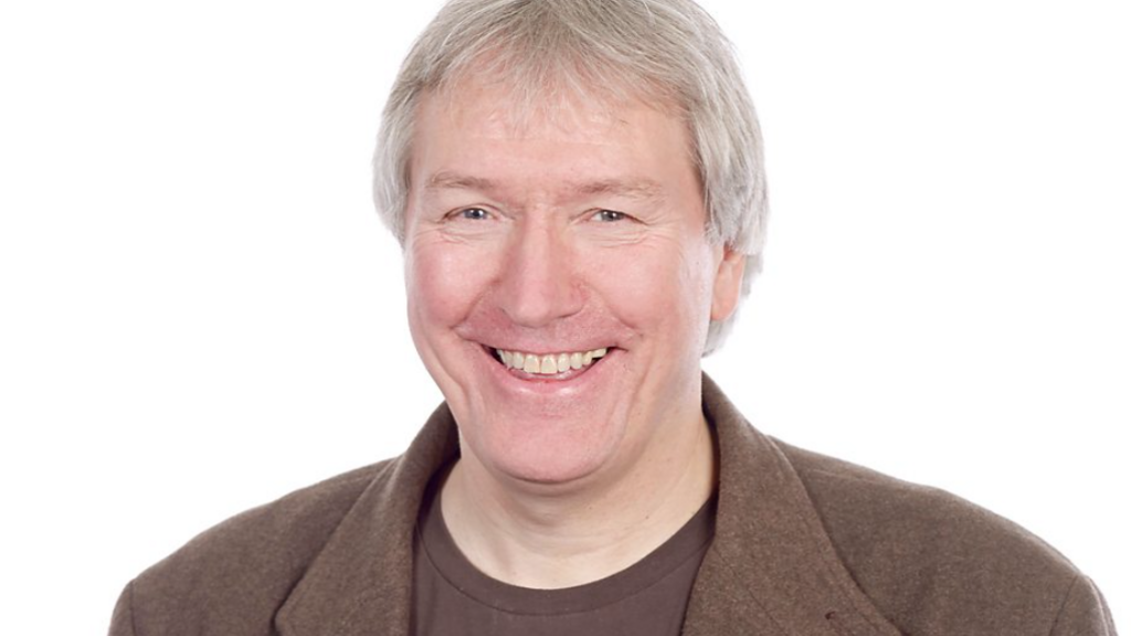 Howard Hughes, a man with grey and white hair smiling at the camera. He is wearing a brown jacket and brown top and is standing against a white background.