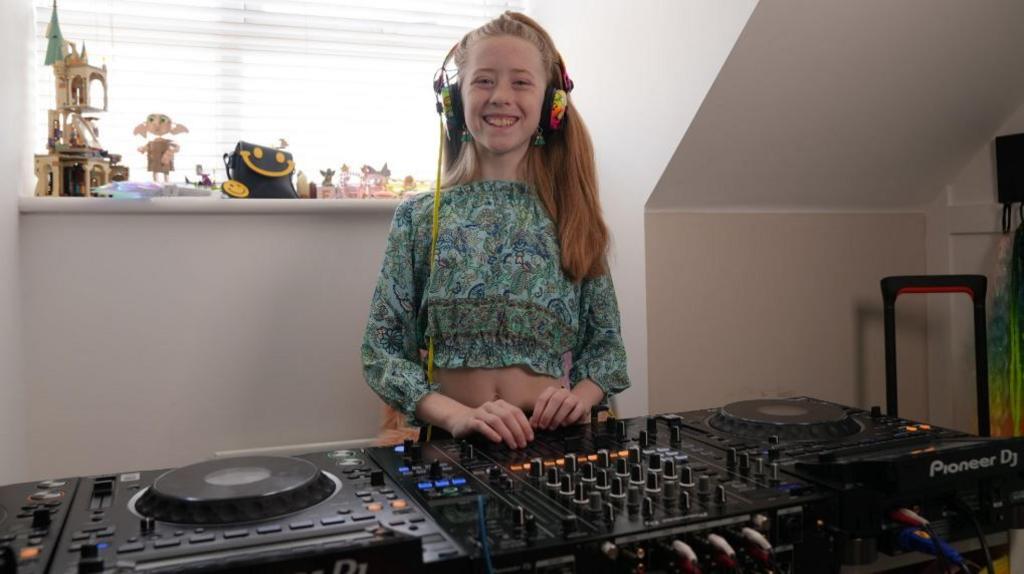 A young girl with long blonde hair is in her house smiling and stood behind some large black DJ decks full of dials and lights.