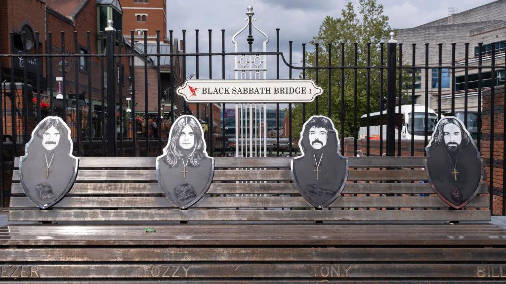 A signpost for Black Sabbath Bridge over a bench featuring the names and metalwork black and white images of members of Black Sabbath on a cast iron black and silver fence overlooking a canal.