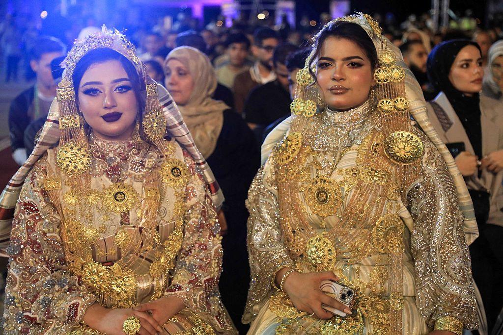 Two women are seated wearing matching golden-coloured dresses and headdresses.