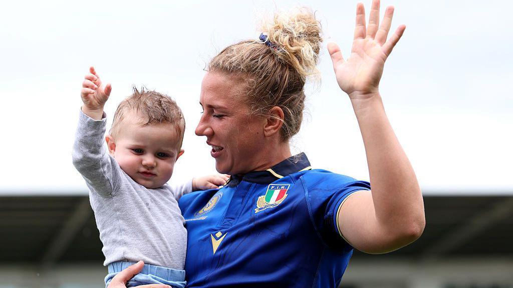 An Italian rugby player in a blue shirt waving to the crowd. She is looking at the very young child in her arm who is also waving.
