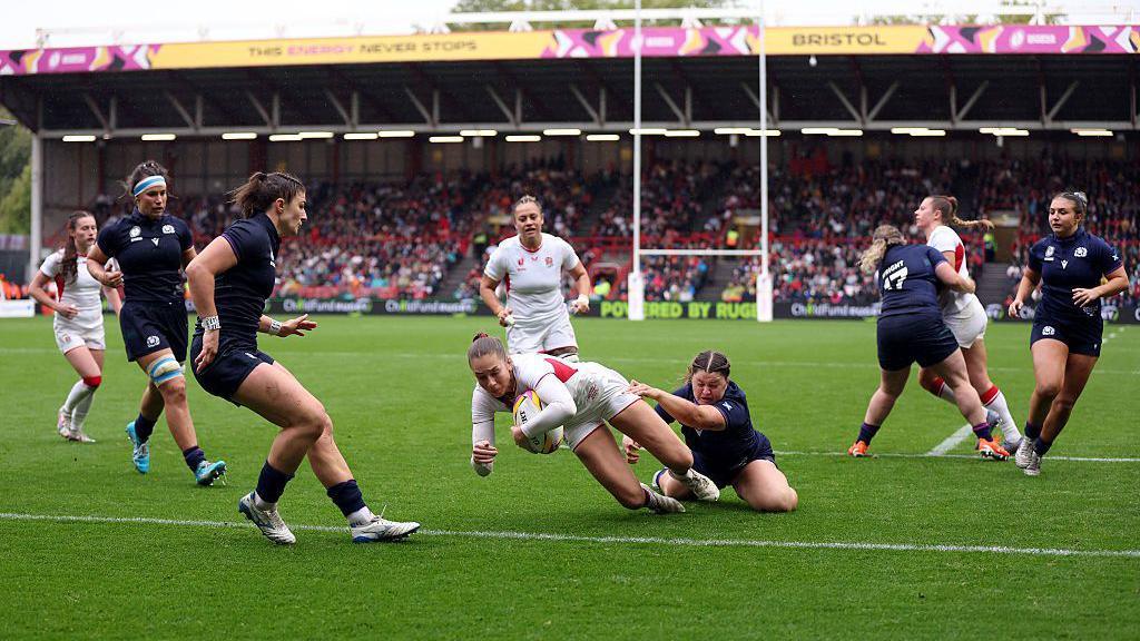 Holly Aitchison of England dives to score her team's sixth try. She is surrounded by Scottish players trying to tackle her.
