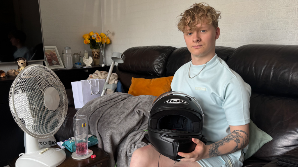 Harry Pyett sitting on a black leather sofa holding his crash helmet. Next to him is a table with a fan and a glass of drink on it. On the left side of the sofa is a blankets and his crutch. He is looking into the camera.
