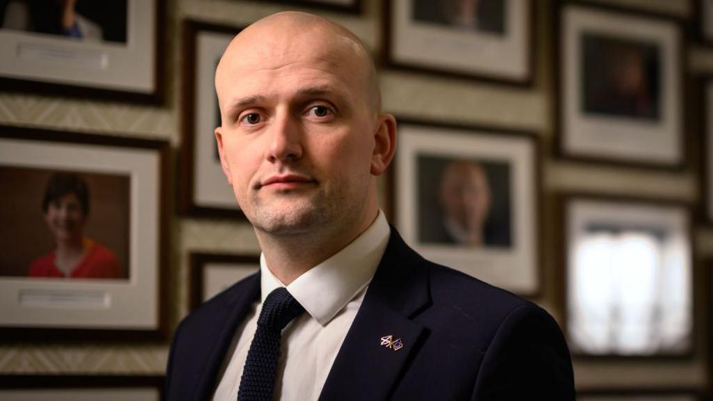 Stephen Flynn, a bald man in a blue suit, blue tie and white shirt, stands in a room full of photos of his SNP colleagues.